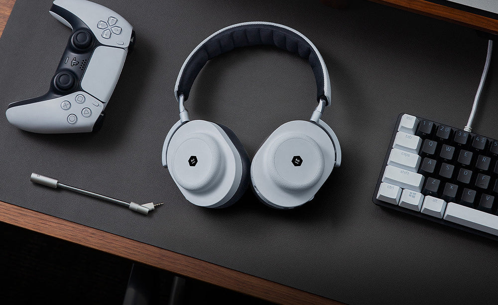 A white headset lying on a table. On the left is a white gaming controller. On the right is a gaming keyboard.